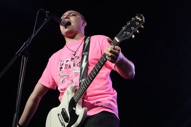 Singer in a pink shirt playing a white guitar onstage.